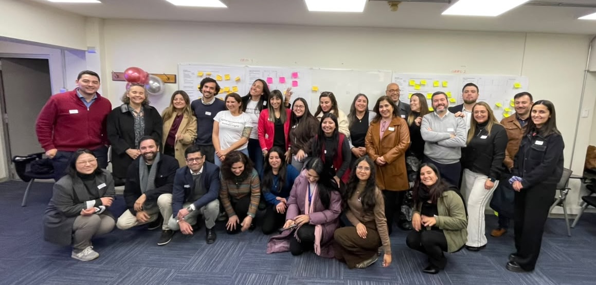 Un grupo de personas, algunas de pie y otras de rodillas, posan juntas en un entorno de oficina frente a pizarras blancas con notas adhesivas.